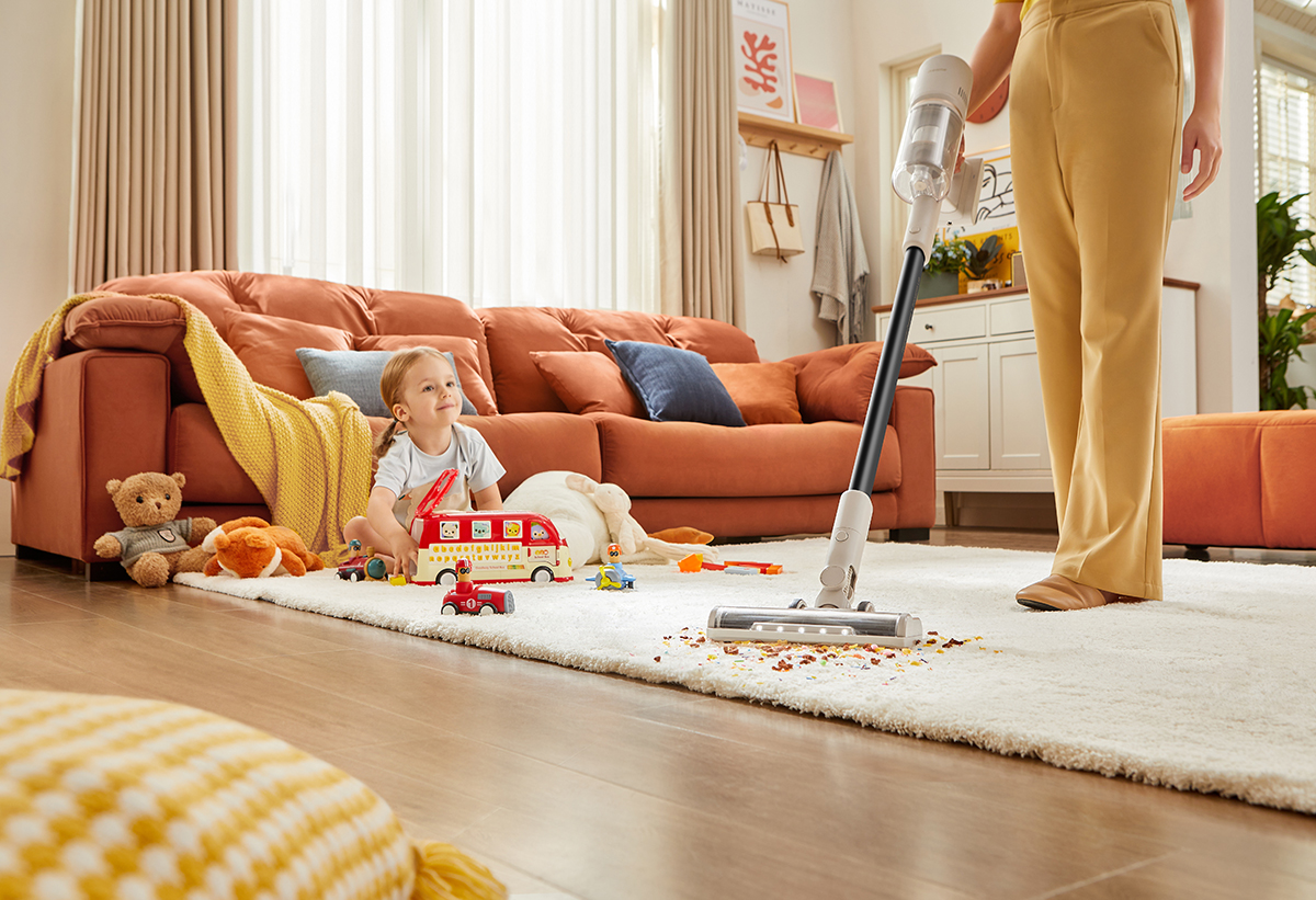 Απεικονίζεται μια γυναίκα να σκουπίζει το χαλί και δίπλα η κόρη της να παίζει με τα παιχνίδια της.